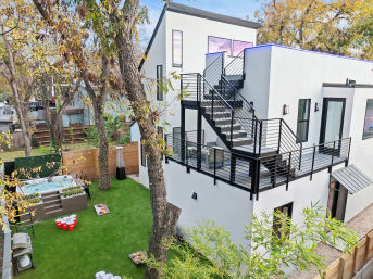 Modern white multi-level home with black metal exterior staircase and rooftop deck overlooking a fenced urban backyard with artificial turf, hot tub, lawn games, and mature trees.