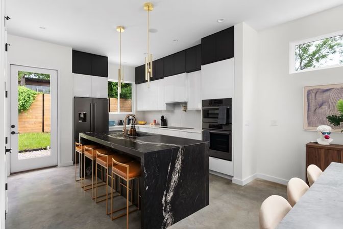 Sleek modern kitchen with a black marble waterfall island, gold pendant lights, four tan leather bar stools, white and black cabinets, stainless appliances, concrete floor and a glass door opening to a fenced backyard.