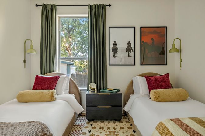 Cozy twin bedroom with two beds flanking a black nightstand, green curtains framing a window with a leafy yard view, olive wall sconces, velvet red and mustard throw pillows, patterned area rug and framed horseback prints on a neutral wall.