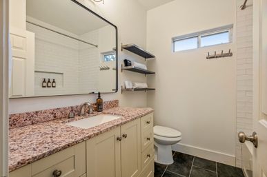 Bright modern bathroom with speckled granite vanity and under-mount sink, chrome faucet and large framed mirror, white subway-tile shower, floating shelves with folded towels and toiletries, small high window, white toilet and dark tile floor — clean spa-like guest bathroom.