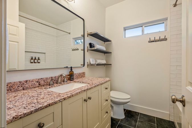 Bright modern bathroom with speckled granite vanity and under-mount sink, chrome faucet and large framed mirror, white subway-tile shower, floating shelves with folded towels and toiletries, small high window, white toilet and dark tile floor — clean spa-like guest bathroom.