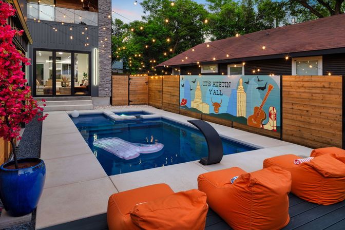 Twilight Austin, Texas backyard with a small rectangular pool and floating lounger, string lights, colorful “It’s Austin Y’all” mural, orange beanbag loungers and a potted bougainvillea