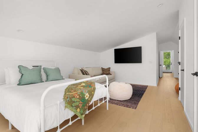 Cozy modern loft bedroom with sloped ceiling, white metal bed with green pillows and floral throw, beige sofa, wall-mounted TV, plush pouf on rug, light oak floors and hallway opening to leafy outdoor view.