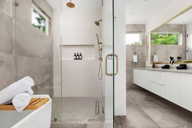 Spa-like modern bathroom with glass walk-in shower and textured white wall, brass rain showerhead and handheld, freestanding tub with wooden tray and rolled white towels, long white floating vanity with large mirror, gray floor tiles and narrow windows showing green trees