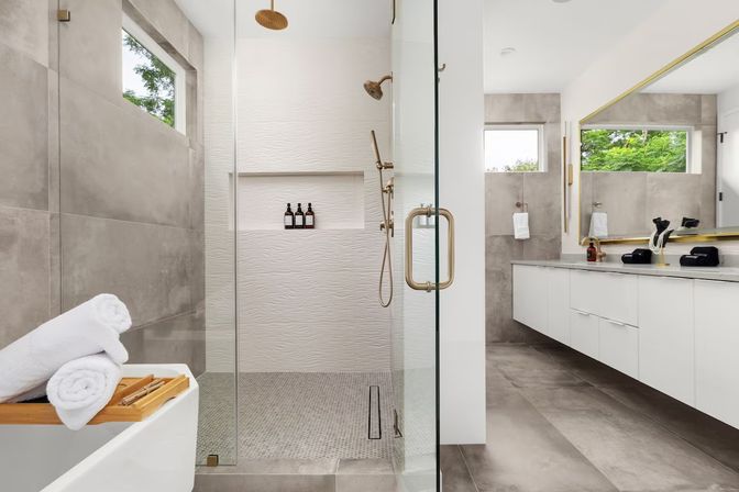 Spa-like modern bathroom with glass walk-in shower and textured white wall, brass rain showerhead and handheld, freestanding tub with wooden tray and rolled white towels, long white floating vanity with large mirror, gray floor tiles and narrow windows showing green trees
