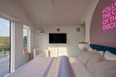 Sunny modern bedroom with balcony view, plush pink bedding and teal headboard, wall-mounted TV, mirrored disco ball and neon sign.