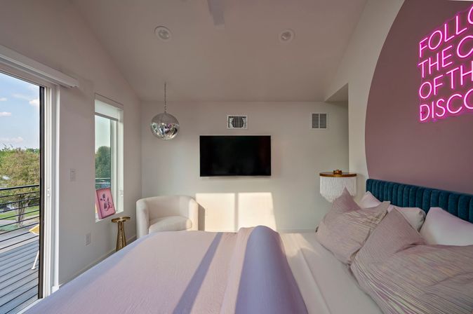 Sunny modern bedroom with balcony view, plush pink bedding and teal headboard, wall-mounted TV, mirrored disco ball and neon sign.