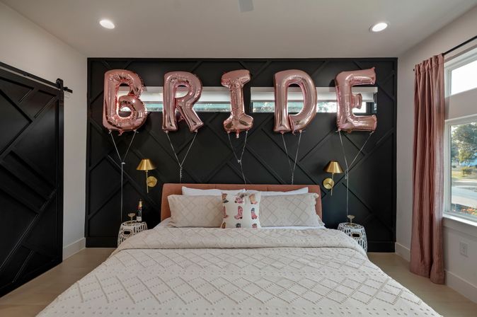 Modern bedroom styled as a bridal suite with rose-gold 'BRIDE' balloons above a blush headboard, white textured quilt, black geometric accent wall, gold wall sconces and natural light from a window.