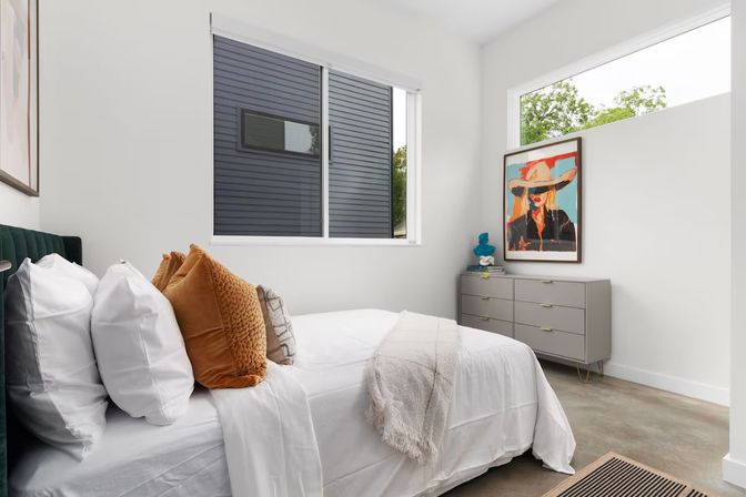 Sunlit contemporary urban bedroom with white walls, low bed dressed in crisp white linens and rust-colored accent pillows, concrete floor, large horizontal windows, and a gray mid-century dresser topped with a colorful pop-art portrait and decor