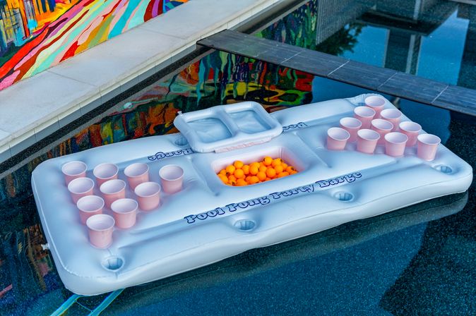 White inflatable beer-pong pool float with pink plastic cups and orange ping-pong balls floating in a backyard swimming pool with a colorful mural reflection.