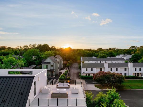 Modern rooftop patio with white outdoor sofas and glass railing, aerial view of suburban townhouses and tree-lined horizon at sunset
