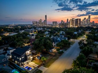 Aerial dusk view of a tree-lined neighborhood and lit modern home with pool, framed by a sunset-glow downtown skyline of high-rise buildings.