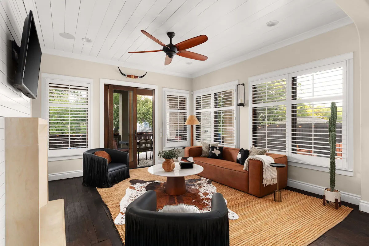 Bright sunroom-style living room with white shiplap ceiling and wooden ceiling fan, brown leather sofa, two black fringe armchairs, round coffee table on jute and cowhide rugs, plantation-shuttered windows and glass doors to a patio, tall potted cactus.