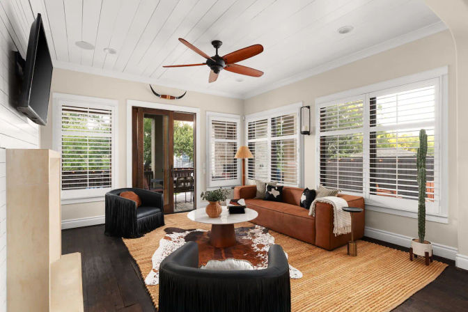 Bright sunroom-style living room with white shiplap ceiling and wooden ceiling fan, brown leather sofa, two black fringe armchairs, round coffee table on jute and cowhide rugs, plantation-shuttered windows and glass doors to a patio, tall potted cactus.