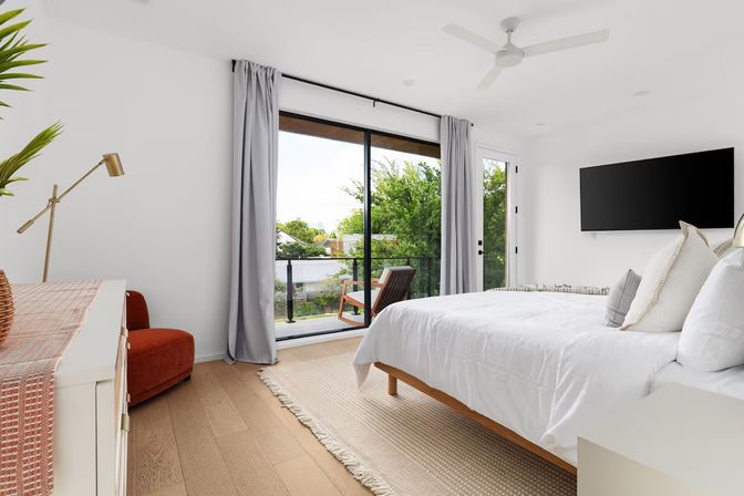 Bright modern bedroom with white linens, wooden platform bed, ceiling fan and wall-mounted TV, sliding glass doors opening to a balcony with a wooden chair and leafy neighborhood view.