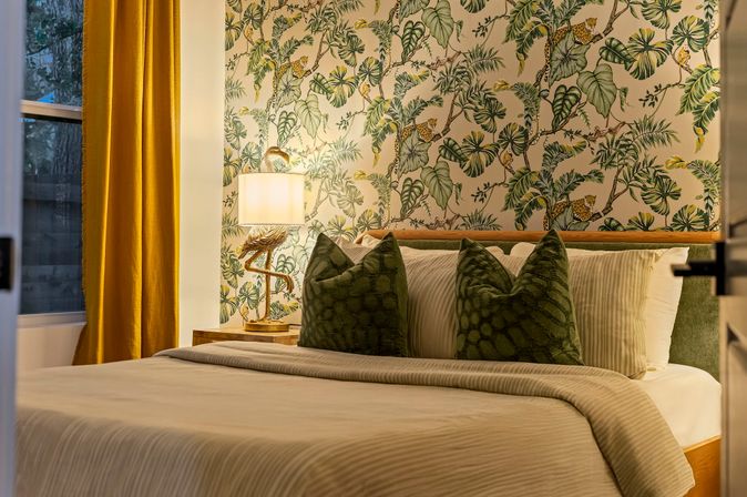 Cozy bedroom with cream bedding, green velvet patterned throw pillows, tropical botanical wallpaper featuring leafy vines and small leopards, brass flamingo table lamp on a wooden nightstand and mustard-gold curtains.