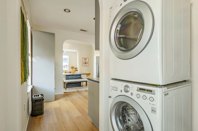 Stacked front-load washer and dryer in a bright, sunlit hallway laundry nook with light wood floors opening to an open-plan kitchen and dining area