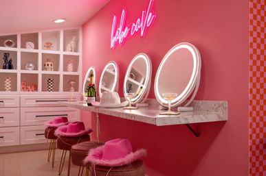 Vibrant pink interior with three illuminated round vanity mirrors above a marble counter, champagne bottle and coupe glasses, fluffy pink cowboy hats on velvet stools, white cubby shelves with decorative accents and a neon cursive sign for a playful, photo-ready makeup station.