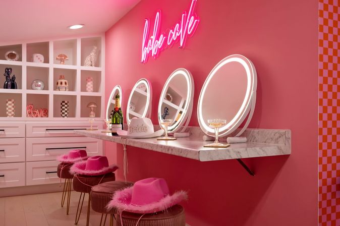 Vibrant pink interior with three illuminated round vanity mirrors above a marble counter, champagne bottle and coupe glasses, fluffy pink cowboy hats on velvet stools, white cubby shelves with decorative accents and a neon cursive sign for a playful, photo-ready makeup station.