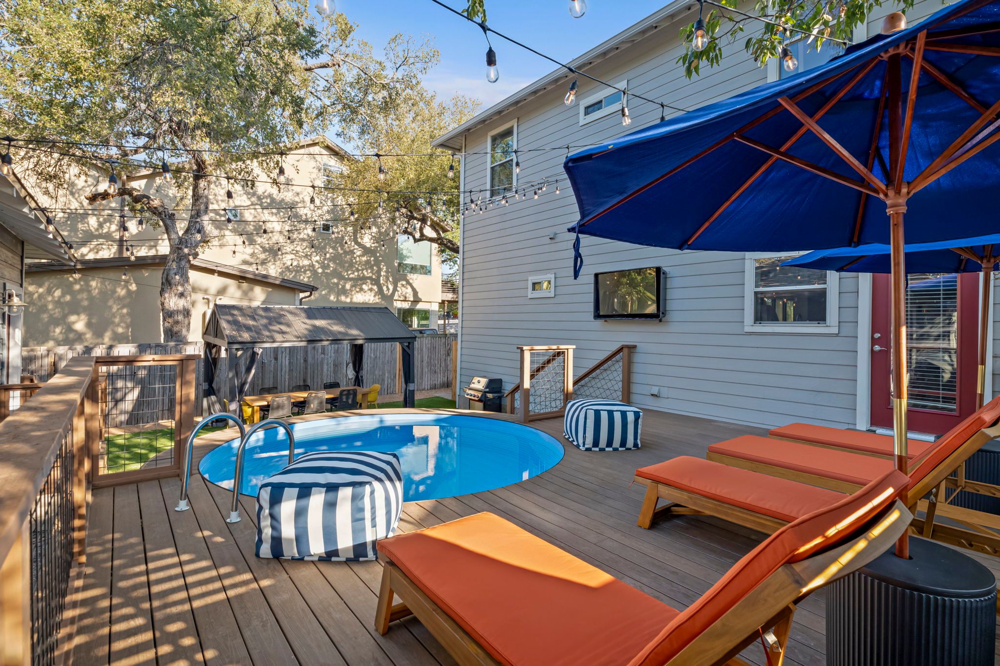 Sunny suburban backyard deck with a round plunge pool, orange-cushioned wooden lounge chairs, blue patio umbrellas, striped poufs, string lights, and a gray two-story house with mature shade trees.