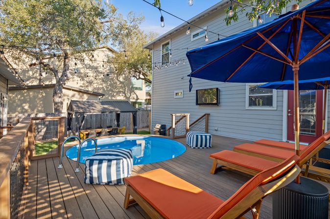 Sunny suburban backyard deck with a round plunge pool, orange-cushioned wooden lounge chairs, blue patio umbrellas, striped poufs, string lights, and a gray two-story house with mature shade trees.
