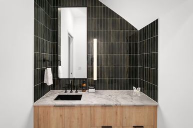 Contemporary home bathroom vanity with dark vertical subway tile accent wall, marble countertop, black undermount sink and matte-black faucet, wood-tone cabinets and vertical LED mirror light.