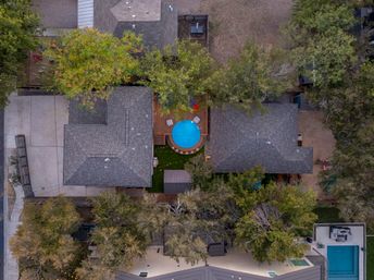 Aerial bird's-eye view of a suburban backyard featuring a round blue above-ground pool on a wooden deck between two houses, surrounded by trees, driveways and neighboring homes.