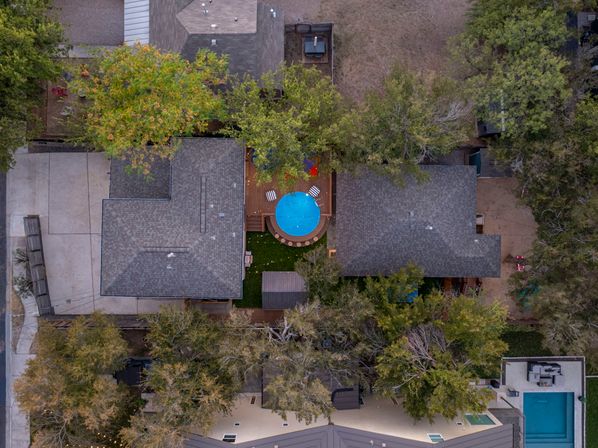 Aerial bird's-eye view of a suburban backyard featuring a round blue above-ground pool on a wooden deck between two houses, surrounded by trees, driveways and neighboring homes.