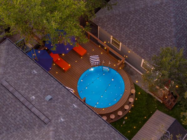 Aerial view of a cozy residential backyard deck at dusk with a round blue above-ground pool lit by string lights, orange lounge chairs, striped ottoman and umbrellas.