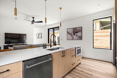 Bright modern open-plan kitchen and living room with a marble-look island, black pull-down faucet, light wood cabinets and floors, stainless dishwasher, brass pendant lights, wall-mounted TV and large window