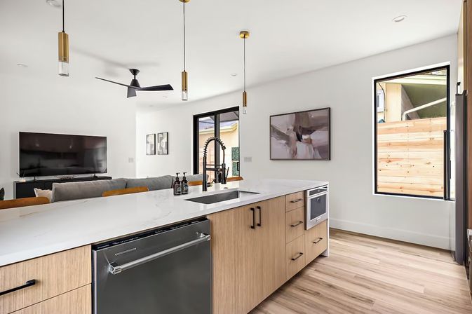 Bright modern open-plan kitchen and living room with a marble-look island, black pull-down faucet, light wood cabinets and floors, stainless dishwasher, brass pendant lights, wall-mounted TV and large window