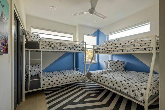 L-shaped kids bedroom with two white metal bunk beds, blue accent walls, black-and-white sunburst-pattern bedding, bold geometric rug, ceiling fan and bright windows