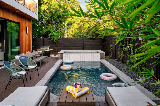 Modern backyard rectangular pool with built-in hot tub, wooden deck, lounge chairs, outdoor dining table and grill, inflatable rings and lush bamboo privacy fence