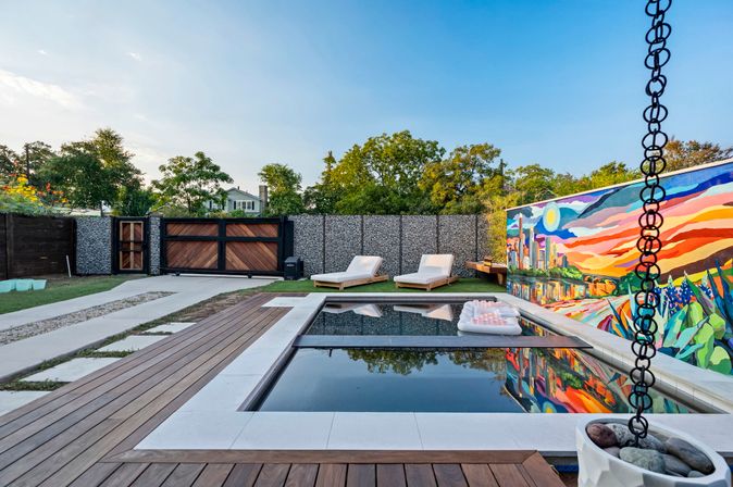 Modern backyard with a rectangular reflecting pool framed by wood decking and white tile, two cushioned lounge chairs on turf, an inflatable pool float, a vibrant cityscape mural on the perimeter wall, and a stone-filled gabion fence with wooden gate.