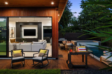 Modern wood-clad backyard deck and pool at dusk with open sliding glass doors to a cozy living room, outdoor dining area, lounge chairs, and a towel cart.