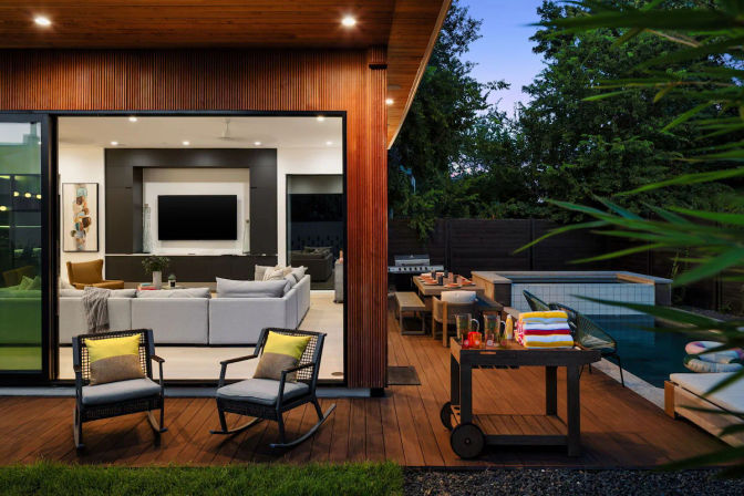 Modern wood-clad backyard deck and pool at dusk with open sliding glass doors to a cozy living room, outdoor dining area, lounge chairs, and a towel cart.