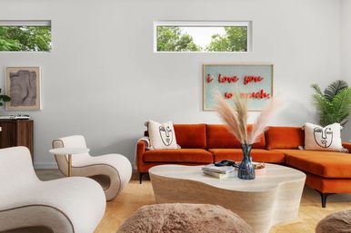 Sunlit modern living room with a burnt-orange sectional sofa, curvy cream lounge chairs, sculptural wooden coffee table topped with a vase of pampas grass, decorative face-print pillows, green plant and wall art reading 'I love you so much'.
