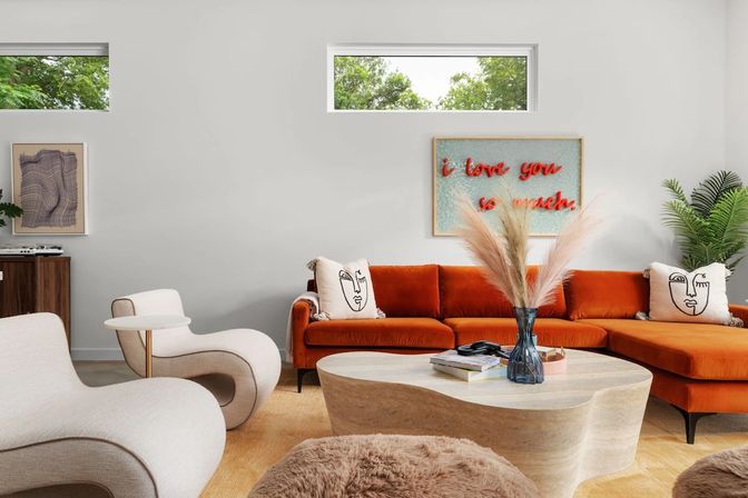 Sunlit modern living room with a burnt-orange sectional sofa, curvy cream lounge chairs, sculptural wooden coffee table topped with a vase of pampas grass, decorative face-print pillows, green plant and wall art reading 'I love you so much'.