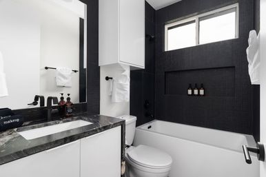 Sleek modern black-and-white bathroom with marble countertop vanity, undermount sink and matte black fixtures, white tub with textured black tile surround and recessed shelf holding amber bottles, wall cabinet, toilet, folded white towels, and frosted window.