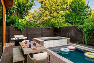 Modern backyard patio with wooden dining table set for eight, stainless grill, raised tiled hot tub and rectangular pool with pastel inflatable rings, dark wood fence and lush trees