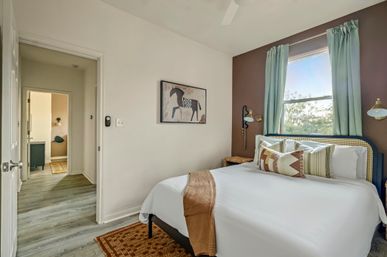 Cozy modern bedroom with white bedding and patterned throw pillows, rattan headboard, teal curtains framing a sunlit window, brown accent wall with horse artwork, wood-look flooring and an open doorway to a hallway.