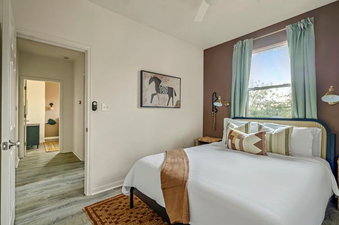 Cozy modern bedroom with white bedding and patterned throw pillows, rattan headboard, teal curtains framing a sunlit window, brown accent wall with horse artwork, wood-look flooring and an open doorway to a hallway.