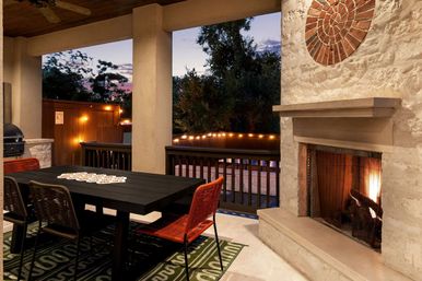 Cozy covered backyard patio at dusk with a lit stone fireplace, black outdoor dining table with orange woven chairs, string lights along a wooden railing and trees beyond.