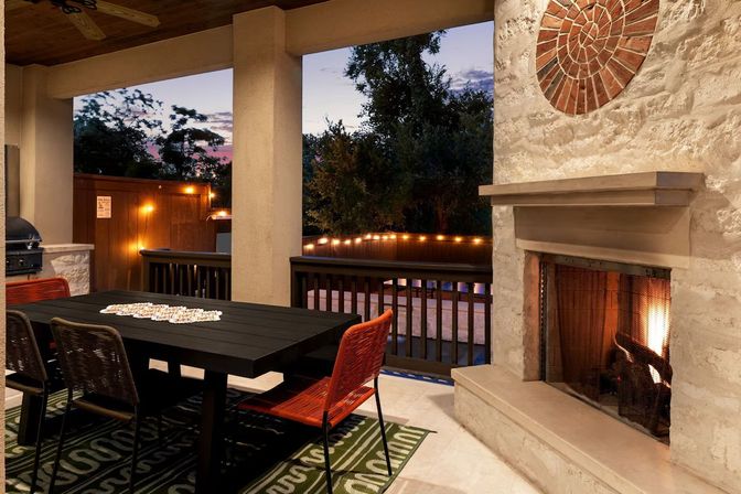 Cozy covered backyard patio at dusk with a lit stone fireplace, black outdoor dining table with orange woven chairs, string lights along a wooden railing and trees beyond.