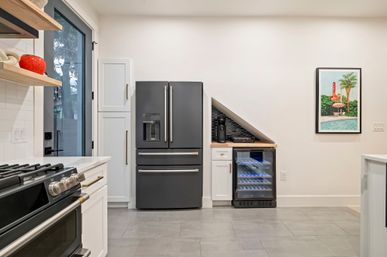 Modern kitchen interior with sleek black French-door refrigerator, gas range, white cabinets, gray tile floor, under-slope wine fridge and coffee station, framed poolside artwork and glass door to yard