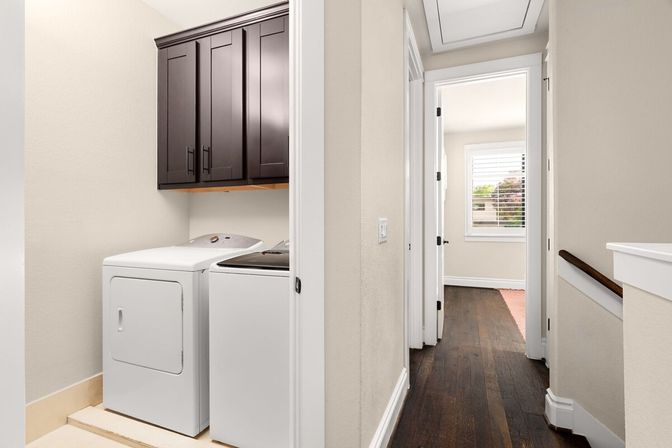 Compact laundry nook with white washer and dryer beneath dark wood cabinets opening to a sunlit hallway with dark hardwood floors and a shuttered window.