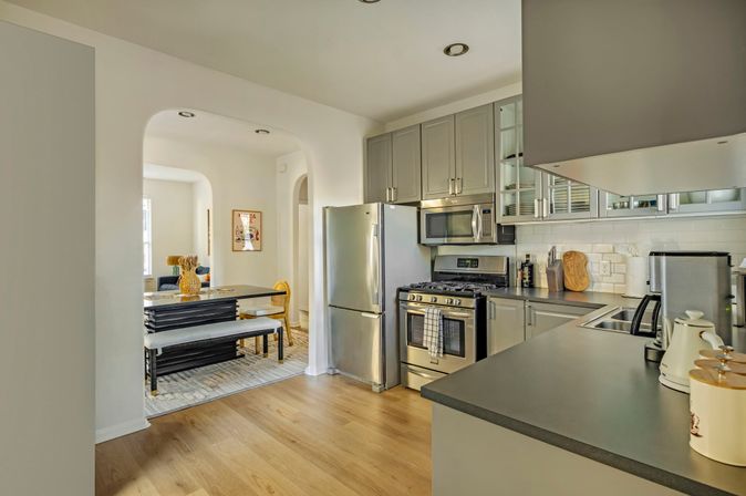 Sunlit open-plan kitchen with stainless steel fridge and gas range, gray cabinets, subway tile backsplash and dark countertops, light wood floors and arched doorway to a dining nook with bench seating and decorative vase.