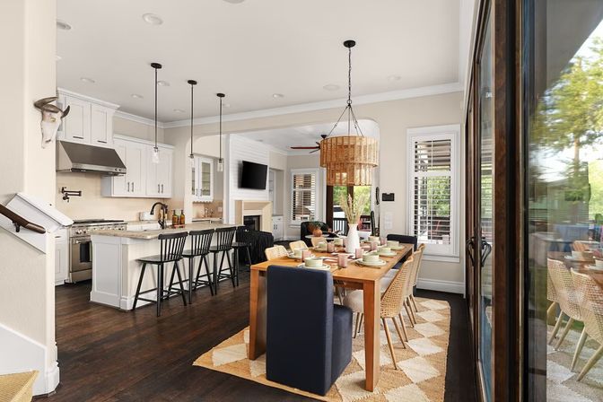 Open-concept kitchen and dining room in a bright suburban home—white cabinets and marble island with black bar stools, wooden dining table set under a woven rattan pendant, dark hardwood floors and sliding glass doors to a backyard.
