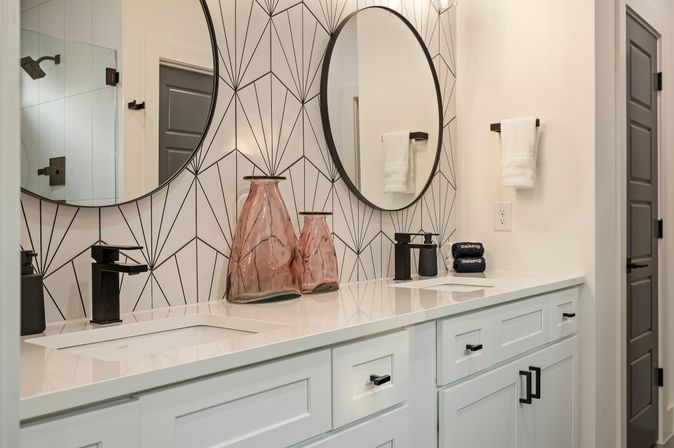Sleek modern double-sink bathroom vanity with white quartz countertop, black faucets, round mirrors, geometric black-and-white tile backsplash and two pink glass vases