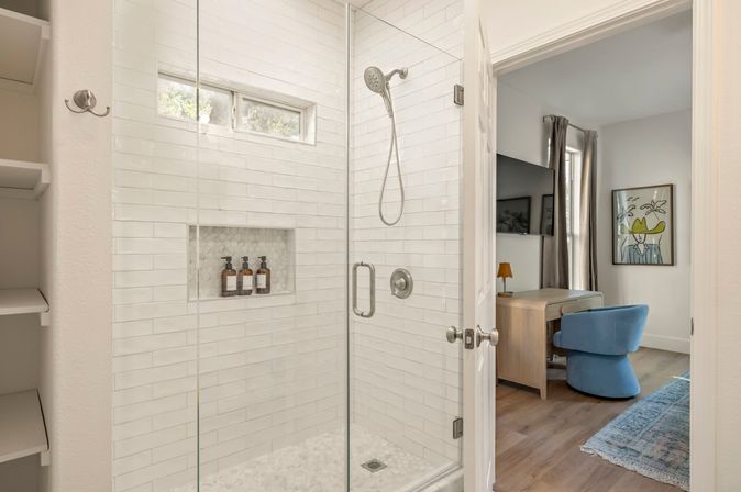 Bright modern frameless glass shower with white subway tile, pebble floor and recessed niche with three pump bottles; open door shows a wood desk, blue swivel chair, wall art and hardwood floors in the adjoining bedroom/office.
